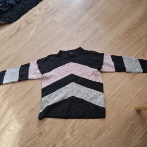 Randig stickad tröja från Vero Moda - Snygg stickad tröja från Vero Moda i storlek S. Tröjan har ett coolt randigt mönster i svart, grått och ljusrosa med breda chevron-ränder. Den har rund hals och långa ärmar, perfekt för att hålla dig varm och stilren under hösten och vintern.