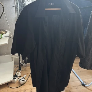 Svart kortärmad skjorta från Mel Richman - Svart skjorta från Mel Richman i storlek XL. Skjortan har klassisk krage, korta ärmar och knäppning framtill. Materialet är en blandning av bomull och polyester, vilket gör den lätt och bekväm. Perfekt för dig som gillar stilrena och enkla plagg.