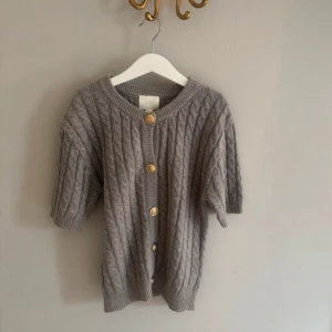 Grå kabelstickad kofta med knappar - Grå kabelstickad kofta med kortare ärmar och snygga guldknappar framtill. Koftan har rund halsringning och ribbade muddar vid ärmslut och nederkant. Perfekt för lager-på-lager och en chill look.