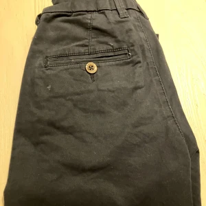 Mörkblåa chinos från Hampton Republic - Snygga mörkblåa chinos från Hampton Republic i storlek 146. Byxorna har klassisk rak passform, bälteshällor och bakficka med knapp. Tillverkade i mjuk bomull som känns skön mot huden. Perfekta för en stilren look.
