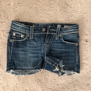 Jeansshorts med broderi från Miss Me - Snygga blå jeansshorts från Miss Me med slitna kanter och dekorativa broderier med nitar på bakfickorna. Shortsen har tydliga kontrastsömmar, dragkedja och coola detaljer på fickorna. Perfekta för dig som gillar en edgy och trendig stil.