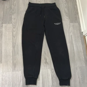 Svarta mjukisbyxor från McKenzie S - Svarta mjukisbyxor från McKenzie med dragsko i midjan och ribbade muddar vid bensluten. Byxorna har en enkel design med vit logga på ena benet och är tillverkade i mjukt material för maximal komfort.