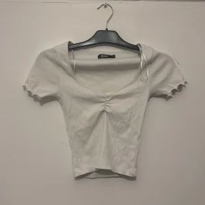 Vit ribbad topp från Bik Bok - Supersöt vit ribbad topp från Bik Bok med korta ärmar och v-ringning. Toppen är croppad och har en snygg rynkdetalj framtill samt vågiga kanter på ärmarna. Perfekt att styla med jeans eller kjol för en fräsch look.
