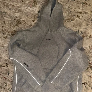 Nike NOCTA grå hoodie med reflexdetaljer - Säljer en grå Nike NOCTA hoodie med snygga reflexdetaljer längs ärmarna och diskret NOCTA-tryck. Klassisk huva med snörning och broderad Nike-logga på bröstet. Mjuk bomullsblandning och ribbade muddar för extra komfort. Perfekt för dig som gillar streetwear. 