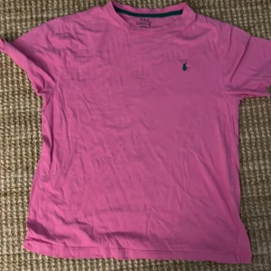 Rosa t-shirt från Polo Ralph Lauren - En klassisk rosa t-shirt från Polo Ralph Lauren med broderad logga på bröstet. T-shirten har rund halsringning och korta ärmar. Perfekt för dig som gillar stilrena och ikoniska plagg med en poppig färg.
