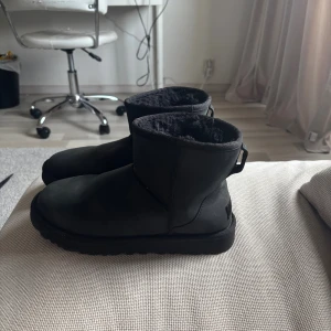 Svarta UGG boots i skinn - Svarta boots från UGG med mjukt foder och klassisk rund tå. Skorna är tillverkade i skinn och har en platt sula samt dragflik baktill. Perfekta för kalla dagar med sin varma insida och stilrena look.
