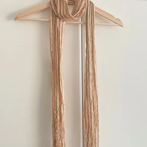 Randig beige och orange mini scarf - Lång och smal halsduk med tunna ränder i beige, orange och bruna toner. Halsduken har en luftig och lätt struktur, perfekt att slänga över axlarna för en avslappnad look. Passar dig som gillar boho-vibbar och mjuka färger.