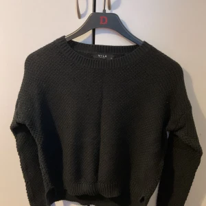 Svart stickad tröja från Vila - Mysig svart stickad tröja från Vila med rund halsringning och lång ärm. Tröjan har en grovstickad struktur och ribbade muddar vid hals, ärmslut och nederkant. Perfekt för kyliga dagar och enkel att matcha med jeans eller kjol.