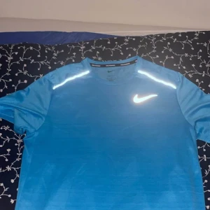 Nike Dri-FIT t-shirt blå & röd bägge 350 st150 - Två Nike Dri-FIT t-shirts i blått och rött med reflekterande detaljer på axlarna och klassisk Nike-logga på bröstet. Perfekta för löpning eller träning, tillverkade i lätt och snabbtorkande polyester. Normal passform och kort ärm.