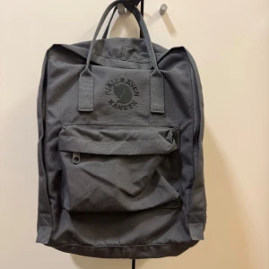 Mörkgrå ryggsäck Fjällräven Kånken - Säljer en klassisk mörkgrå Fjällräven Kånken ryggsäck med broderad logga framtill. Ryggsäcken har ett stort huvudfack, en praktisk framficka med dragkedja och två sidofickor. Perfekt för skola, jobb eller utflykter.