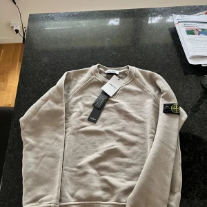 Beige sweatshirt från Stone Island Junior - Säljer en beige sweatshirt från Stone Island Junior med klassisk rund hals och ikonisk logotyp-patch på ärmen. Tröjan är i mjuk bomull och har ribbade muddar vid ärmslut och nederkant. Perfekt för dig som gillar stilrena och exklusiva plagg.