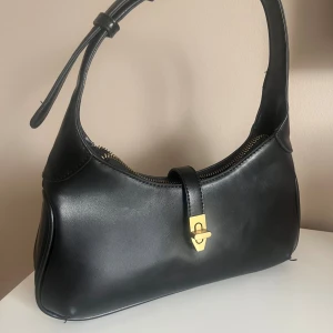 Svart axelväska i skinn med guldspänne - Stilren svart axelväska i slätt skinn med justerbart axelband och snygga gulddetaljer. Väskan har dragkedja upptill och ett elegant spänne framtill. Perfekt storlek för det viktigaste och ger en clean look till din outfit.