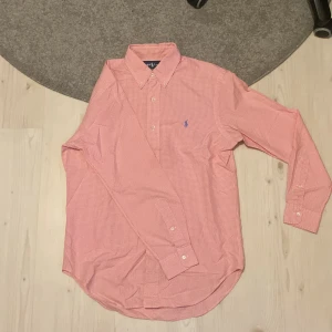 Rutig rosa skjorta från Ralph Lauren - Säljer en snygg rosa och vit rutig skjorta från Ralph Lauren med klassisk passform. Skjortan har lång ärm, knappar framtill och blå broderad logga på bröstet. Perfekt för dig som gillar stilrena och tidlösa plagg.