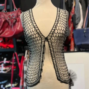 Svart och silver nätväst med pärlor - Unik nätväst i svart med silverfärgade pärlor och detaljer. Västen har v-ringad front, knäppning med små bollar och ett öppet, transparent mönster. 