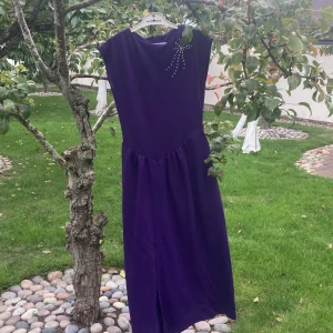 Lila långklänning med pärldetalj - Långklänning i djup lila med ärmlös design och markerad midja. Klänningen har en snygg pärldetalj på axeln och stängs med dragkedja i ryggen. Tillverkad i polyester, perfekt för dig som vill sticka ut med en elegant och stilren look.