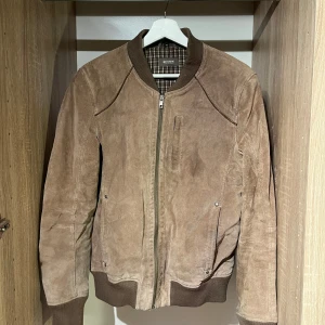 Brun mocka bomberjacka  - brun bomberjacka i mjuk mocka från M.Town. Jackan har ribbade muddar vid krage, ärmslut och nederkant, samt dragkedja framtill. Insidan har ett rutigt foder. Satt pris ex frakt