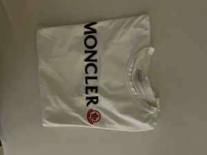 Vit Moncler t-shirt med tryck - Snygg vit t-shirt från Moncler i storlek M med stort svart Moncler-tryck vertikalt på framsidan och röd logga. Klassisk rund hals och korta ärmar. Mjuk och skön bomullskvalitet som känns lyxig och stilren.