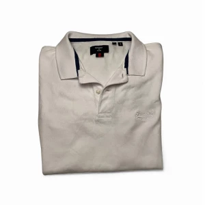 Vit Superdry piké  - Vit pikétröja från Superdry med klassisk krage och knappar framtill. Tröjan har diskret broderad logga på bröstet och mörkblå detalj på insidan av kragen. Tillverkad i bomull för en skön och luftig känsla. Sitter jättebra och har inga defekter överhuvudtaget. Storlek S