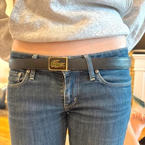 Svart skärp från Lacoste med gulddetalj - Snyggt svart skärp från Lacoste med en rektangulär spänne i guld och svart, prydd med den klassiska krokodilloggan. Skärpet är gjort i slätt skinn och har flera hål för justering. Perfekt accessoar för att lyfta din outfit.