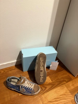 Philippe Model blå-bruna sneakers - Snygga sneakers från Philippe Model med en cool mix av blå denim och bruna mockadetaljer. Skorna har vit snörning, rund tå och platt sula med tydlig logga undertill. Perfekt för dig som gillar streetstyle och vill sticka ut med ett unikt par.