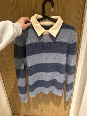 Randig ullpiké från Polo Ralph Lauren - Säljer en klassisk pikétröja från Polo Ralph Lauren i 100% ull. Tröjan har breda marinblå och ljusblå ränder, krage i beige och knäppning framtill. Perfekt för dig som gillar preppy stil och vill ha något både snyggt och bekvämt.