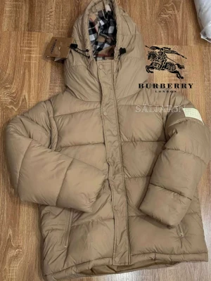 Beige Burberry Puffer - Säljer en elegant och mångsidig Burberry-jacka i svart, i nyskick. Dunfodrad för värme och komfort med det klassiska rutmönstret i huvan som ger en exklusiv touch. Praktiska dragkedjefickor, avtagbar huva och diskret logopatch på ärmen. Passar M/L. Jackan är i princip som ny och utan defekter. En lyxig kombination av stil och funktion – perfekt för både vardag och tillfällen då du vill klä upp dig.  📦Snabb leverans - skickas samma dag📌 (om ej ev hindr.)