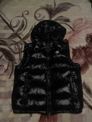 Svart dunväst från Moncler - Snygg svart dunväst från Moncler med glansig finish och huva. Västen har två framfickor med dragkedja och klassisk Moncler-logga på bröstet. Insidan har coolt serietryck och märkning. Perfekt för att ge din outfit en lyxig touch.