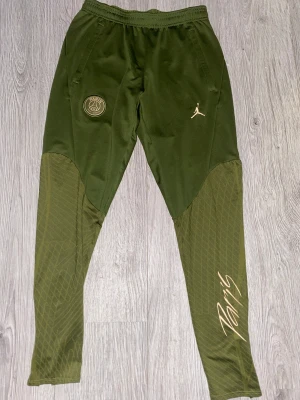 Jordan PSG gröna slim fit träningsbyxor - Snygga gröna träningsbyxor från Jordan PSG med slim fit-passform. Byxorna har elastisk midja, Paris Saint-Germain-logga, guldfärgad Jumpman och Paris-tryck på benet. Materialet är polyester med elastan och benen har diskret mönster för extra stil.