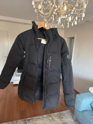 Svart Sail Racing dunjacka med huva - Svart dunjacka från Sail Racing med huva och snygga logodetaljer på bröstet och ärmen. Jackan har lång ärm, dragkedja framtill och quiltad design som ger en cool och sportig look. Perfekt för kalla dagar när du vill hålla stilen.