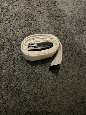 Vit canvasbälte från Urban Classics - Snyggt och stilrent vitt canvasbälte från Urban Classics med svart metallspänne där loggan är ingraverad. Bältet har en klassisk rak form och är tillverkat i slitstark bomull med detaljer i svart metall. Perfekt för att ge din outfit en clean och modern touch.