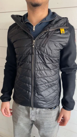Parajumper cardigan  - Svart pufferjacka från Parajumpers med huva och hel dragkedja framtill. Jackan har quiltad front och rygg i glansigt material, samt släta, stickade ärmar. Detaljer som gul knapp vid halsen och logopatch på axeln. Perfekt för kyliga dagar.