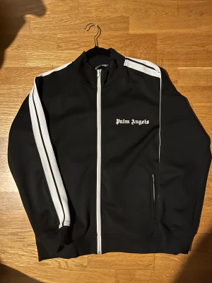 Svart vindjacka Palm Angels - Palm Angels svart vindjacka med vita ränder längs ärmarna och vit dragkedja. Jackan har ståkrage, lång ärm och logotypen broderad på bröstet. Snygg och sportig design som sticker ut med kontrastdetaljer.