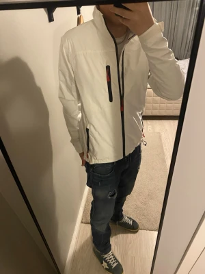 Vit vindjacka Helly Hansen S - Snygg vit vindjacka från Helly Hansen med svarta detaljer och röda dragkedjor. Jackan har hög krage, tre fickor med dragkedja och meshfoder på insidan. HH-logga på ärm och rygg, samt justerbar nederkant. Perfekt för dig som gillar sportig stil.