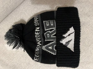 Svart och grå Åre mössa med tofs - Stickad svart och grå mössa med stor tofs i svart, grått och vitt. Texten 'ÅRE' och 'SWEDEN' syns framtill samt ett broderat bergmotiv på uppviket. Perfekt för kalla dagar och vintersäsongen.