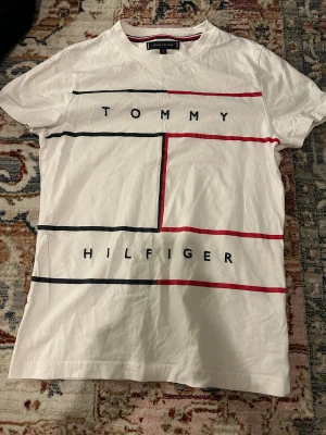 Vit Tommy Hilfiger t-shirt XS - Vit t-shirt från Tommy Hilfiger i storlek XS med grafiskt tryck i svart och rött på framsidan. Klassisk rund hals och korta ärmar. Materialet är mjuk bomull och designen har en modern, stilren vibe med tydlig logga.