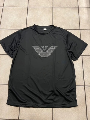 Svart t-shirt med stor logga, XL - Svart t-shirt i 100% polyester med en stor, stilren logga framtill i silver. Klassisk rund hals och korta ärmar. Perfekt för dig som gillar en sportig och clean look.