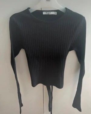 Svart ribbad tröja med öppning i ryggen - Svart långärmad topp från NA-KD med ribbad struktur och öppning i ryggen med knyt. Toppen har en croppad passform och rund halsringning. Perfekt för dig som gillar stilrena och trendiga plagg med snygga detaljer