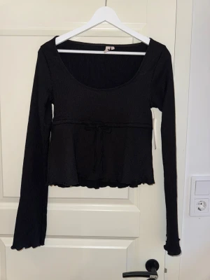 Svart ribbad långärmad topp med knyt - Svart långärmad topp från Nelly med ribbad struktur och söt knytning under bysten. Modellen har en rundad, djup urringning och volangkant vid ärmslut och nederkant. Perfekt att styla med jeans eller kjol för en chill look.