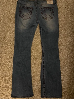 True Religion blå bootcut jeans - Säljer ett par blå jeans från True Religion med klassiska röda sömmar och ikoniska fickor bak. Modellen har låg midja och bootcut-ben, vilket ger en snygg siluett. Jeansen är tillverkade i jeansmaterial och har detaljerade fickor med knappar framtill.