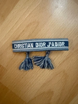 Blå Christian Dior J'Adior armband - Snyggt blått armband från Christian Dior med texten 'J'Adior' broderad i vitt. Armbandet har två tofsar i ändarna och ett randigt mönster längs kanterna. Perfekt accessoar för att lyfta din outfit med lite lyxig vibe.
