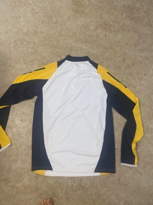 Real Madrid träningströja Adidas - Säljer en snygg Real Madrid träningströja från Adidas med lång ärm. Tröjan är vit med marinblå och gula paneler, har halv dragkedja och klubbmärke på bröstet. Perfekt för dig som älskar fotboll och vill ha en cool look på träningen.