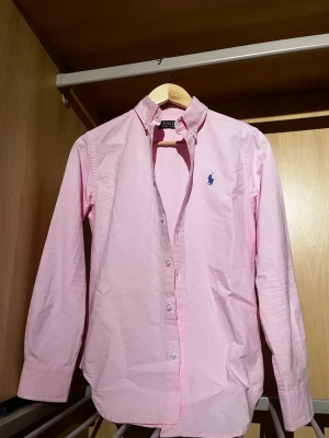 Rosa skjorta från Polo Ralph Lauren - Snygg rosa skjorta från Polo Ralph Lauren med klassisk button-down krage och blå broderad logga på bröstet. Skjortan har långa ärmar, knappar framtill och är gjord i mjuk bomull. Perfekt för dig som gillar stilrena och tidlösa plagg.