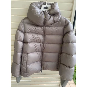 Beige pufferjacka från Add - Säljer en beige pufferjacka från Add med hög krage och dragkedja framtill. Jackan är tillverkad i ultralätt nylon med vattenavvisande yta och har ett satinliknande tyg. Fylld med dun för extra värme och har ribbade muddar vid ärmslut. Perfekt för kalla dagar.