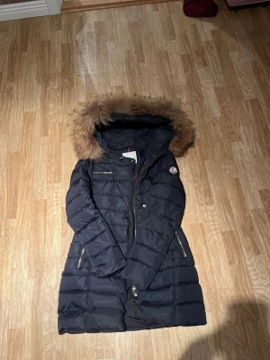 Svart Moncler dunjacka - Snygg svart dunjacka från Moncler med pälskantad huva och quiltad design. En liten del av dragkedje knappen har försvunnit (synd på bilden) men det påverkar inget!