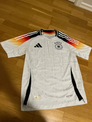 Tyskland Tröja - Säljer en officiell Tyskland fotbollströja från Adidas. Tröjan är vit med svarta detaljer och har färgglada mönster i rött, orange och gult på axlarna. Klassisk Adidas-logga och landslagsemblem på bröstet. Perfekt för fotbollsfans!