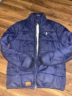 Mörkblå pufferjacka från Polo Ralph Lauren - Snygg mörkblå pufferjacka från Polo Ralph Lauren med klassisk broderad logga på bröstet. Jackan har dragkedja framtill, högt krage och två sidofickor med zip. Materialet är glansig polyester och jackan har quiltade paneler för extra värme. Jackan passar mig bra och jag är 1,87 och den passar mig bra