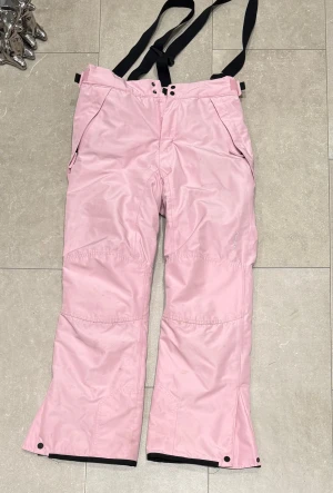 Rosa täckbyxor med hängslen, storlek 164 - Säljer ett par ljusrosa täckbyxor med svarta justerbara hängslen. Byxorna har två fickor med dragkedja, förstärkta knän och knappar vid benslut. Perfekta för vintern och håller dig varm och torr under kalla dagar.