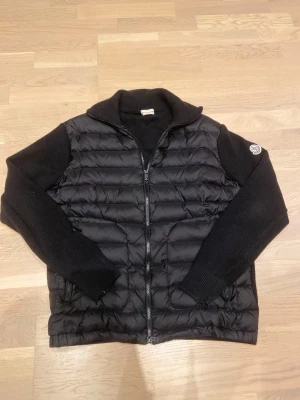 Svart dunväst från Moncler - Svart Cardigan från Moncler med quiltad front och stickade svarta ärmar. Cardiganen har dragkedja framtill, hög krage och Moncler-logga på vänster ärm. Materialmix av dun och stickad polyester ger en schysst kontrast och modern look. Skriv gärna för fler bilder eller frågor !🤩