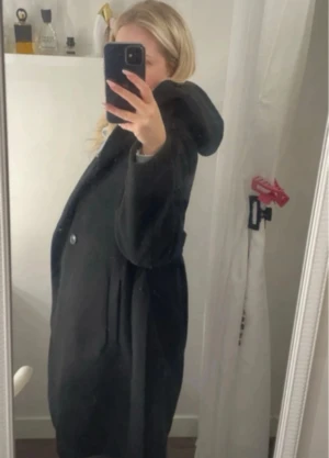 Svart kappa med huva - Säljer en svart kappa med huva och knappar framtill. Jackan har en loose passform och är perfekt för kyliga dagar. Materialet känns mjukt och värmande, och den har en stilren look som funkar till många outfits.