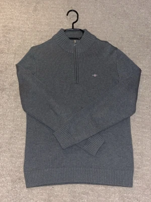 Grå stickad half zip tröja GANT - Snygg mörkgrå stickad tröja från GANT med half zip och ribbad krage. Liten broderad logga på bröstet. Perfekt för dig som gillar klassisk stil med en modern touch. Långärmad och tillverkad i mjuk bomull för skön komfort.